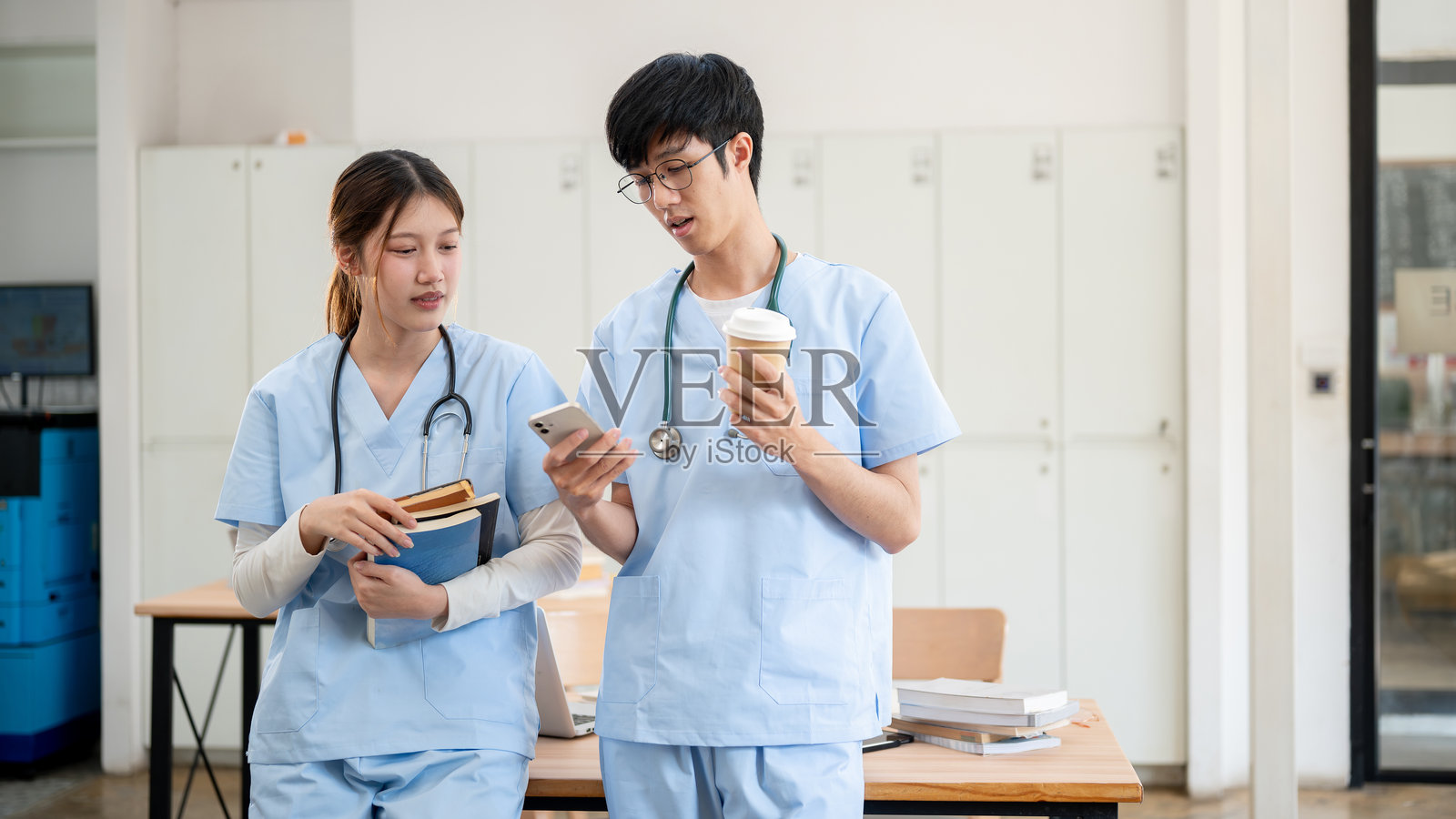 两名快乐的亚洲医学生穿着医务服,站在教室里讨论。照片摄影图片