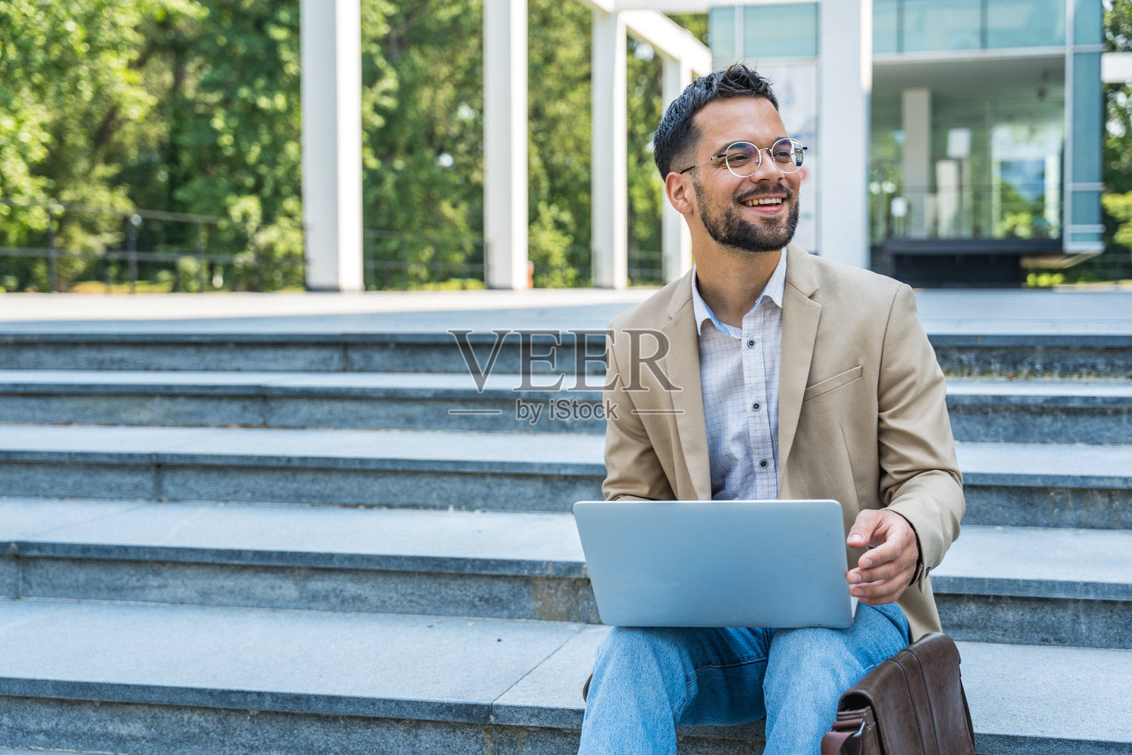 快乐聪明的男性商人企业家,专业职业,正在使用笔记本电脑。商人在办公室大楼外坐着,利用笔记本电脑工作于新项目。照片摄影图片