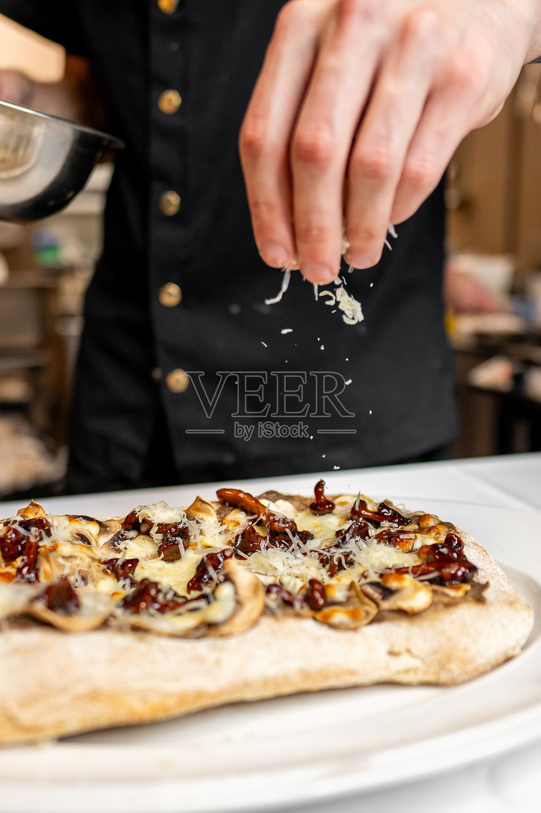 一位厨师在现代厨房中将磨碎的奶酪撒在顶有蘑菇和焦糖洋葱的美味扁面包上,展示了其烹饪技巧。照片摄影图片