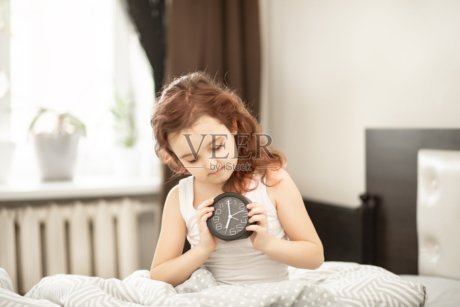 可爱的小女孩坐在床上,早晨醒来,手里拿着闹钟。照片摄影图片