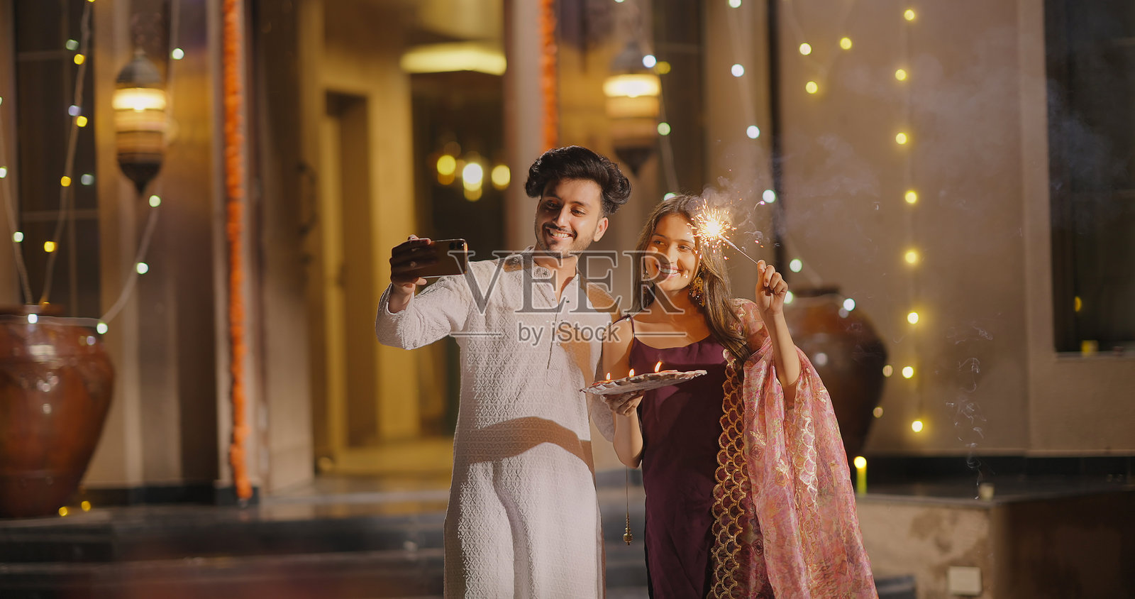 印度教徒快乐的Z世代情侣在户外家中摆出可爱的姿势自拍,享受愉快的时刻。年轻的丈夫和妻子手持手机,面向镜头,享受着灯火辉煌的排灯节夜晚装饰。照片摄影图片