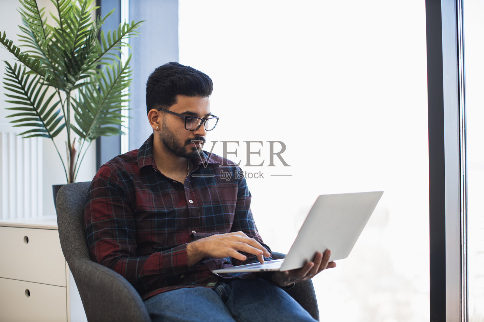 年轻的印度男子在大窗户旁使用笔记本电脑工作照片摄影图片