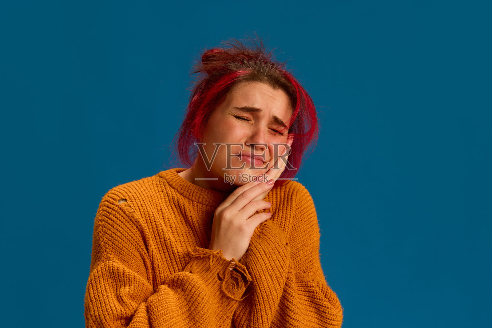 女人捂着脸颊,表现出牙痛,常因病毒炎症或免疫力下降而加重,背景为蓝色工作室。照片摄影图片