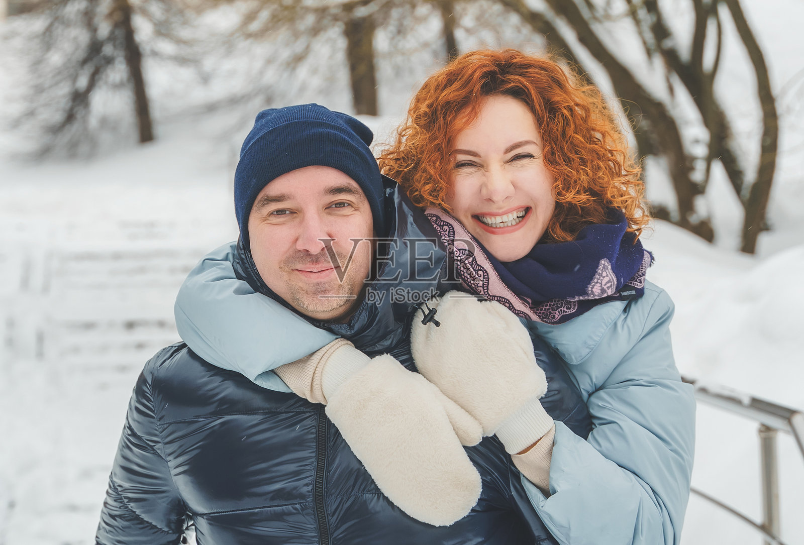 浪漫爱情故事。快乐的中年千禧夫妇,冬季雪地公园里一位红发女子。浪漫约会,亲吻,拥抱。散步,享受乐趣。时尚的蓝色衣服,夹克,帽子。照片摄影图片