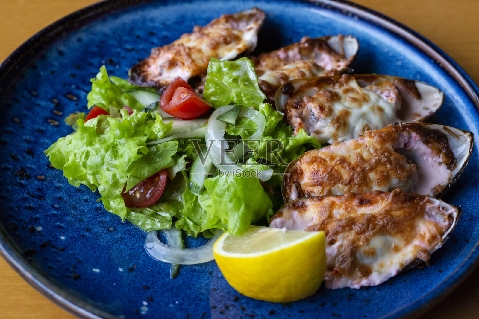 海鲜。贝类贻贝。烤贻贝配奶酪、香菜和柠檬,放在铸铁黑盘上。贻贝盘,冷白葡萄酒,柠檬和香菜,背景为旧蓝色木板。照片摄影图片