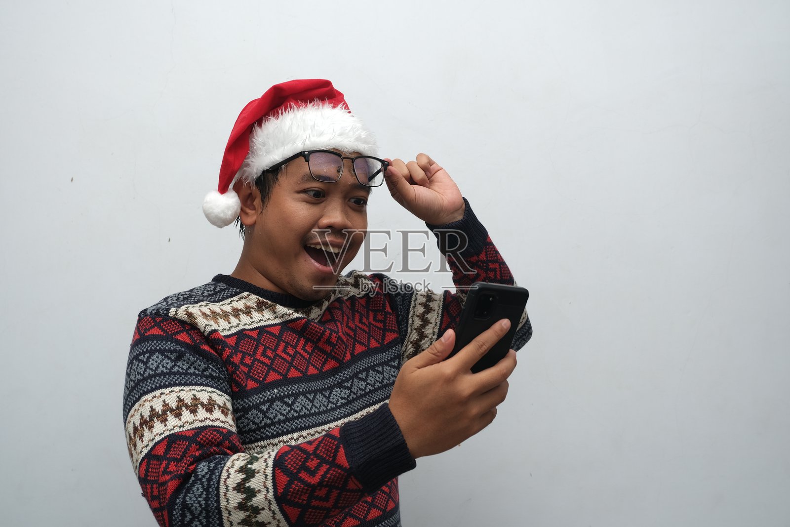 兴奋的亚洲男子穿着节日毛衣和圣诞老人帽,惊讶地看着手机并调整眼镜。照片摄影图片