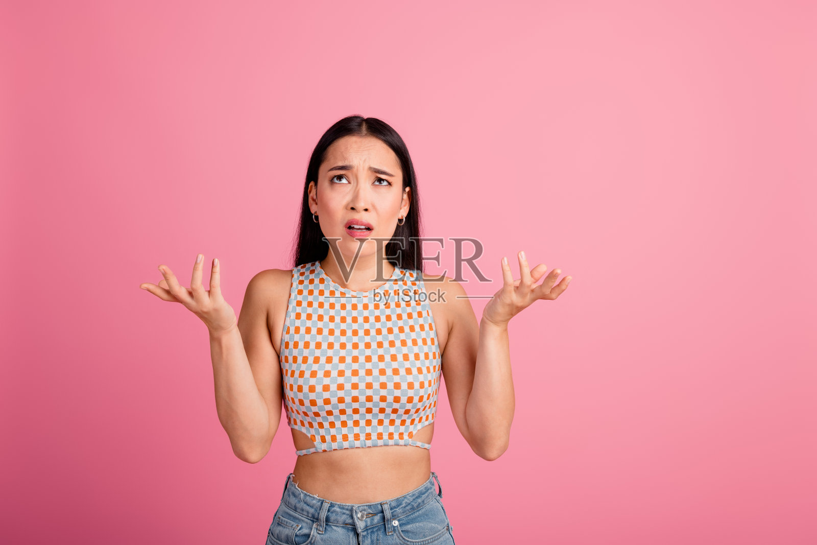 困惑的年轻女性,长棕色头发,站在粉色背景前,双臂举起,表达不确定感。照片摄影图片