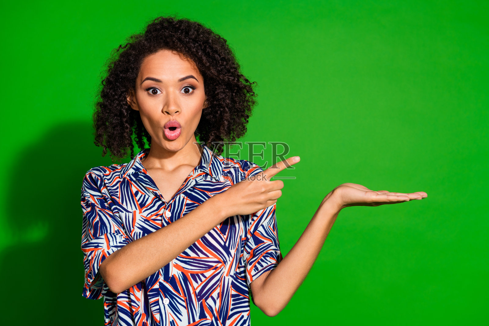 迷人的女士穿着印花衬衫,指着空旷的手臂,背景为绿色。照片摄影图片