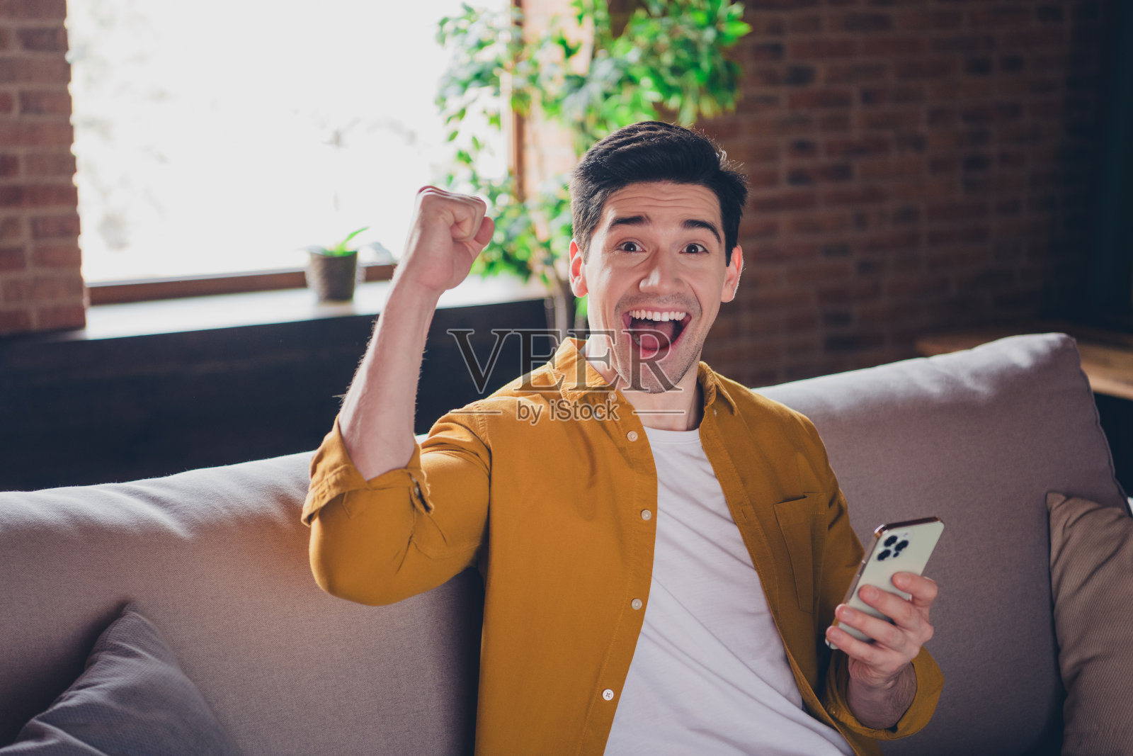 兴奋的年轻人在时尚的阁楼公寓里庆祝成功,使用智能手机,阳光透过窗户洒入室内。照片摄影图片