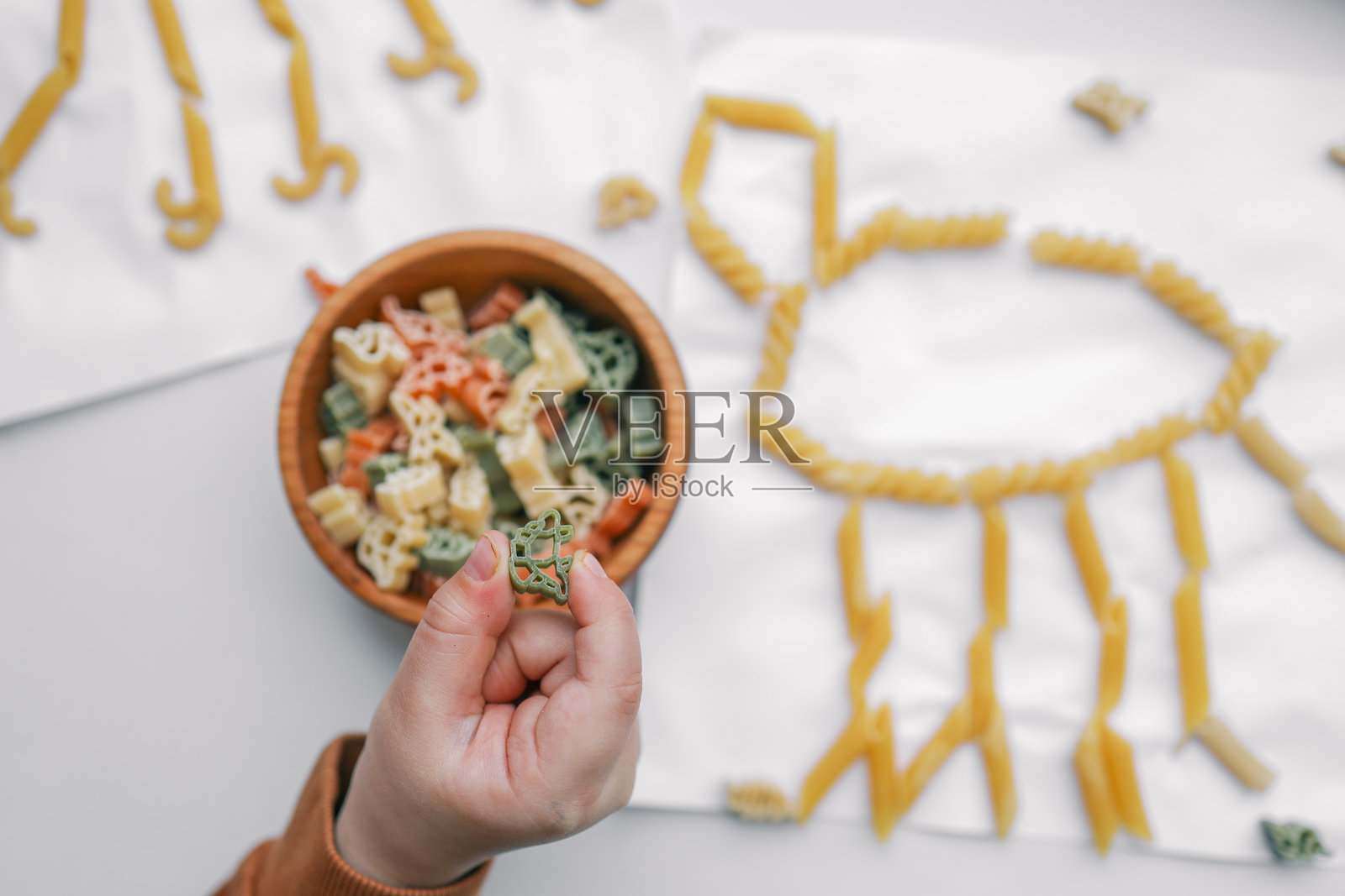 孩子们的创意手工活动,使用意大利面艺术在白纸上形成动物形状。一只小手握着五颜六色的意大利面,旁边有一个装有各种意大利面片的碗。适合儿童的有趣DIY艺术项目。照片摄影图片