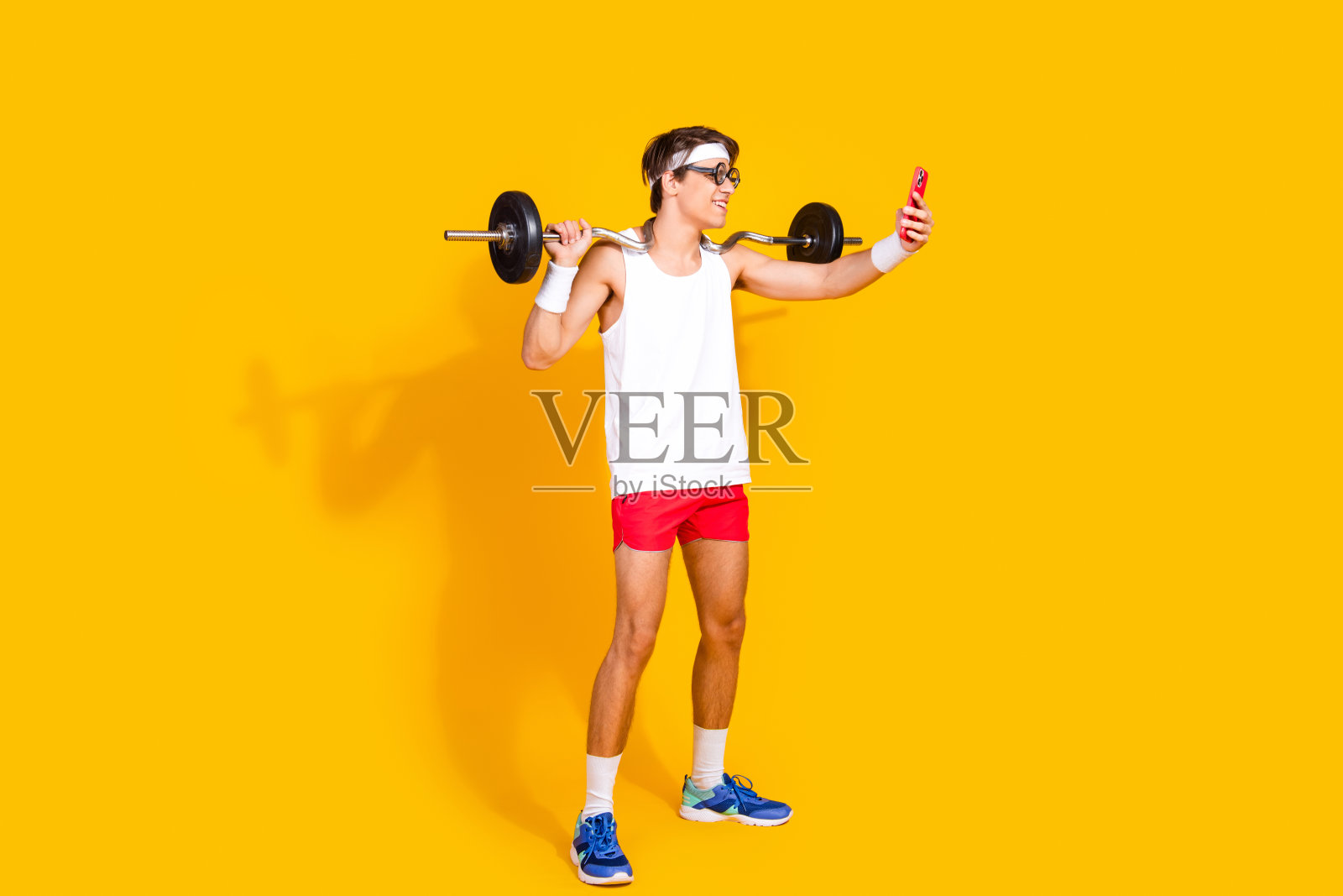 年轻男子穿着运动服在鲜艳的黄色背景下举着杠铃自拍。照片摄影图片