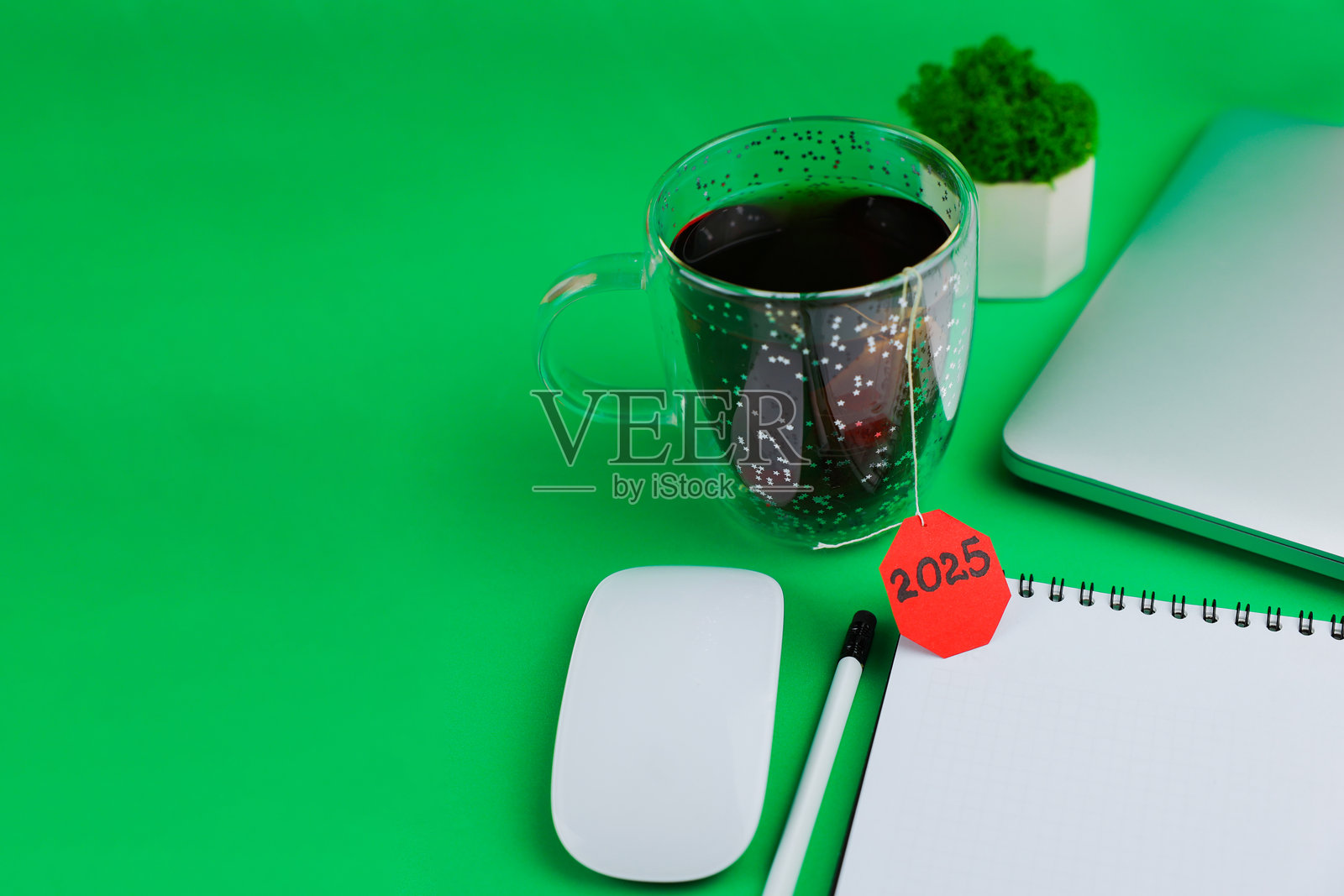 从上方和侧面看一个透明茶杯,杯子上有一个鲜红色的标签,上面写着2025,杯子放在明亮的绿色背景上,周围有白色的钞票和一支白色铅笔,可以看到笔记本电脑的边缘和一块清洁的绿色布。照片摄影图片