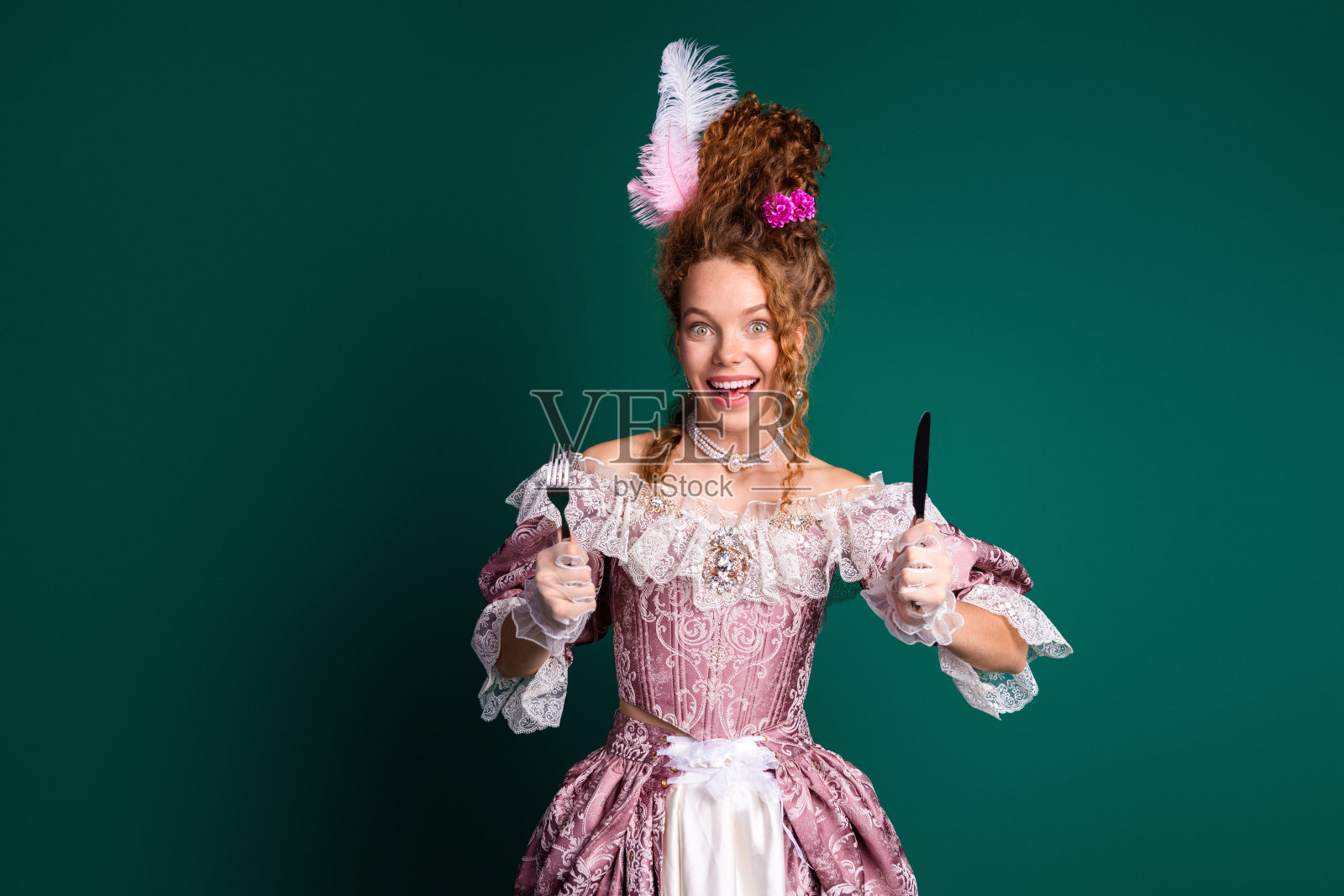 穿着巴洛克风格粉色裙子的快乐年轻女性在绿色背景前手持餐具的肖像照片摄影图片