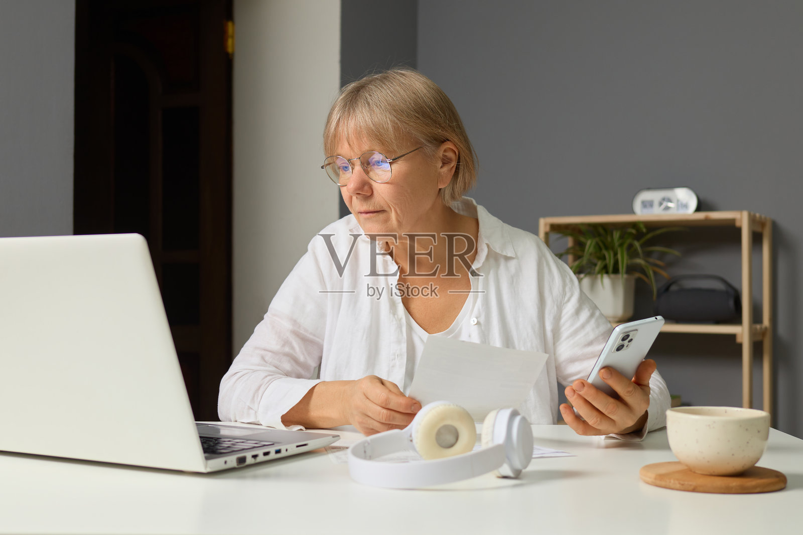 一位年长女性专注于她的工作,坐在整洁的桌子前,使用笔记本电脑并握着智能手机。她似乎在审阅文件并整理任务,营造出一个舒适的工作环境。照片摄影图片