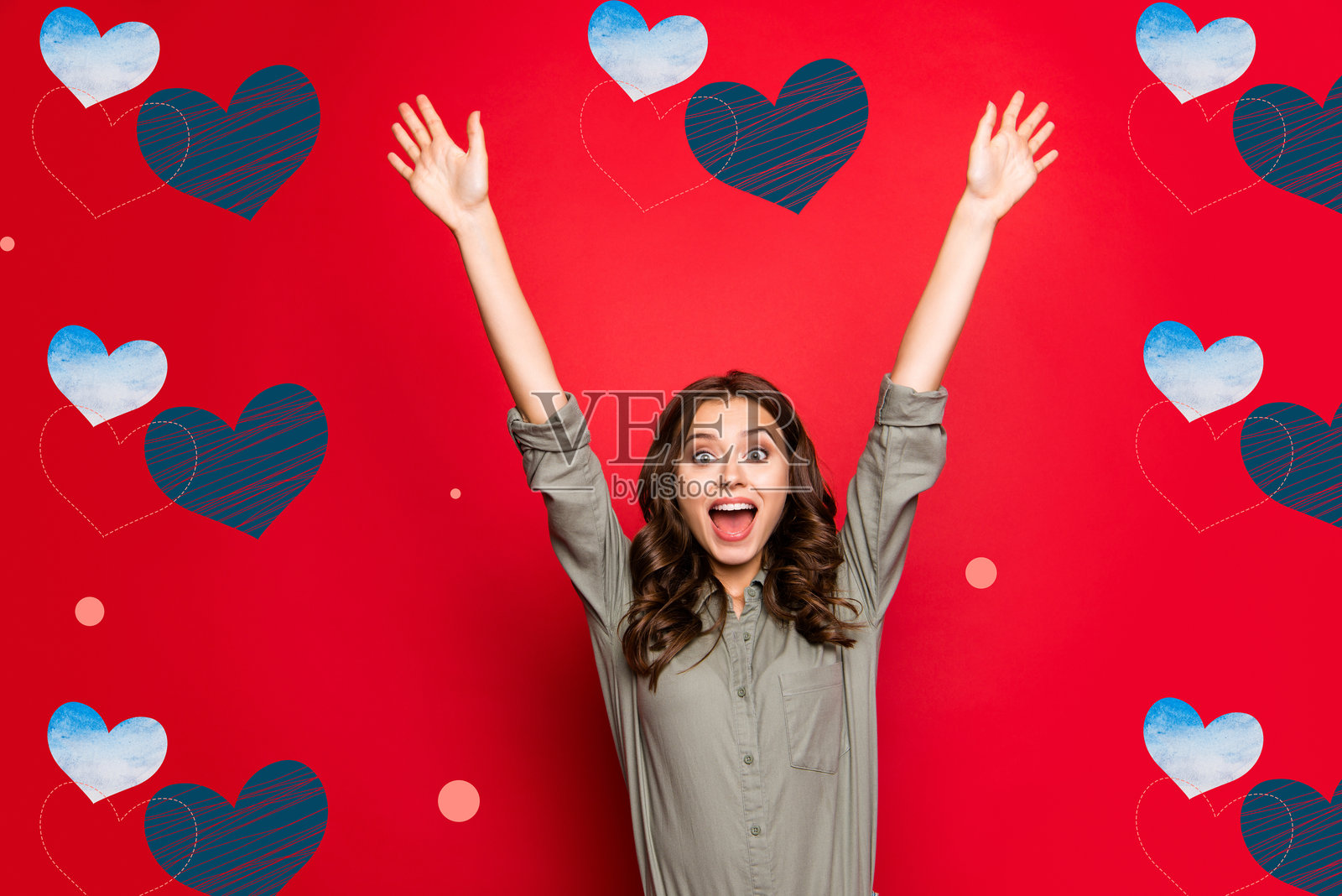 一位穿着衬衫的快乐、无忧无虑的女士,棕色现代卷发,双手举起,张嘴微笑,背景为鲜艳的红色。照片摄影图片