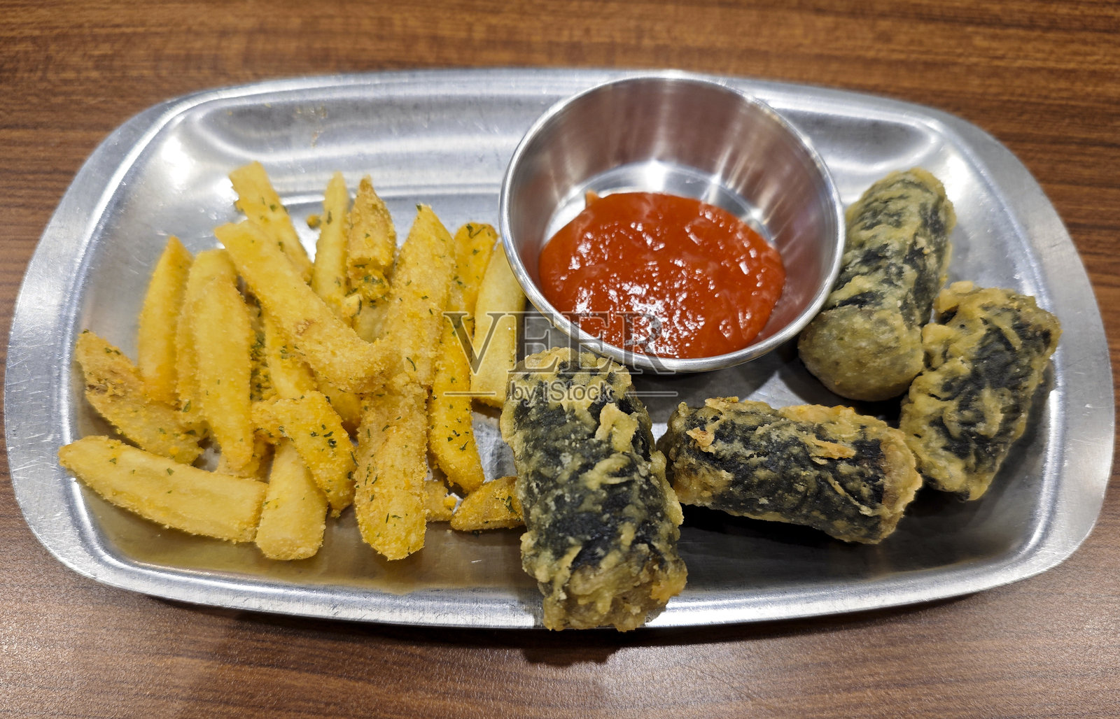 一盘脆皮韩国炸海苔卷和调味法式薯条,配有辛辣红色蘸酱,放在银色托盘上,置于木桌上。照片摄影图片