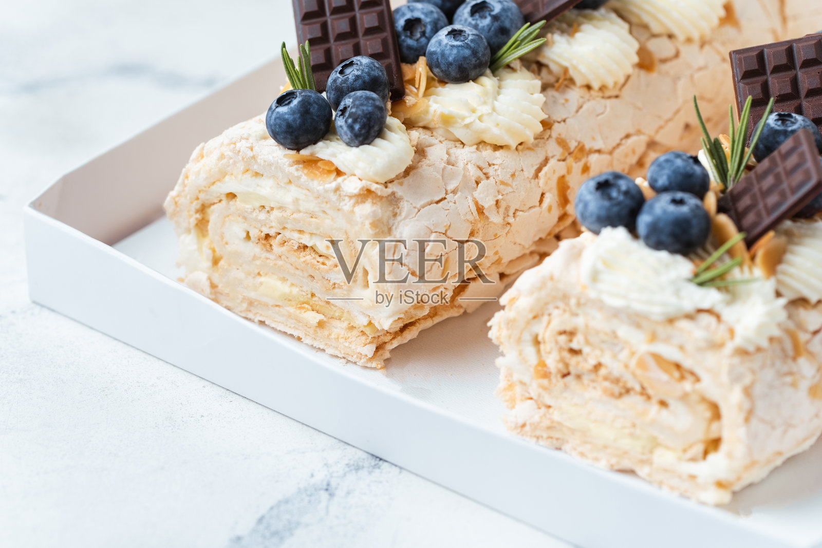 自制的蛋白霜卷,装饰有杏仁片、蓝莓和迷你巧克力棒,放在白色礼品盒中,背景为白色。蛋白霜卷顶部覆盖着鲜奶油。照片摄影图片