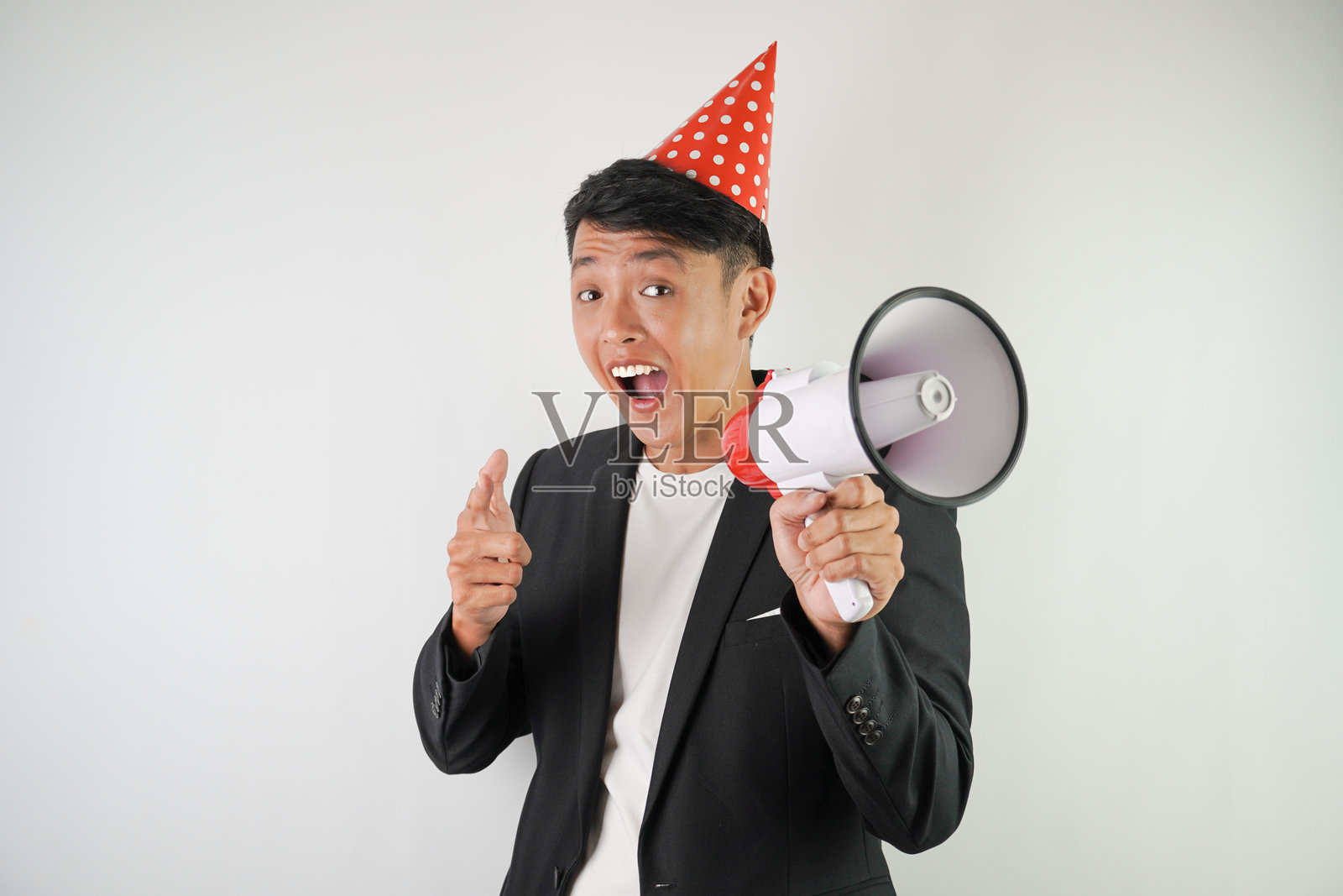 兴奋的年轻亚洲男子戴着红色派对帽,穿着白衬衫和黑色西装,正在用扩音器大喊,背景为白色。适用于圣诞节和新年派对的概念。照片摄影图片