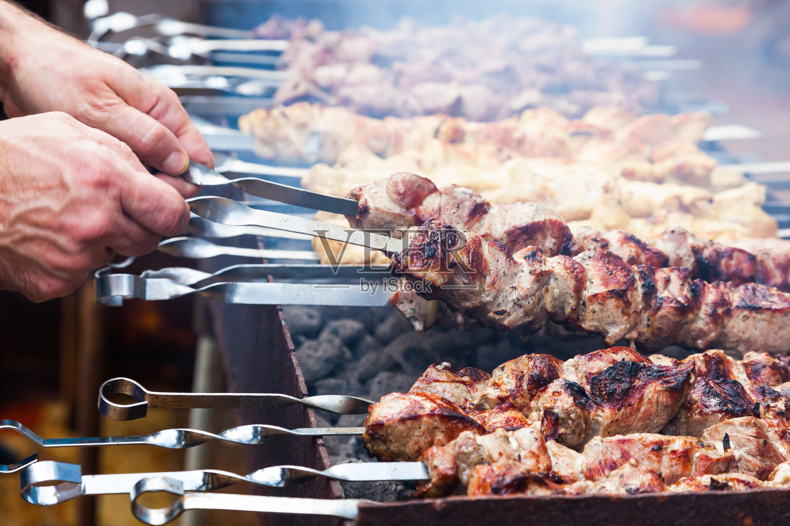 男人的手正在烤架上煎烤串。烤架上有猪肉、鸡肉和羊肉的串。照片摄影图片