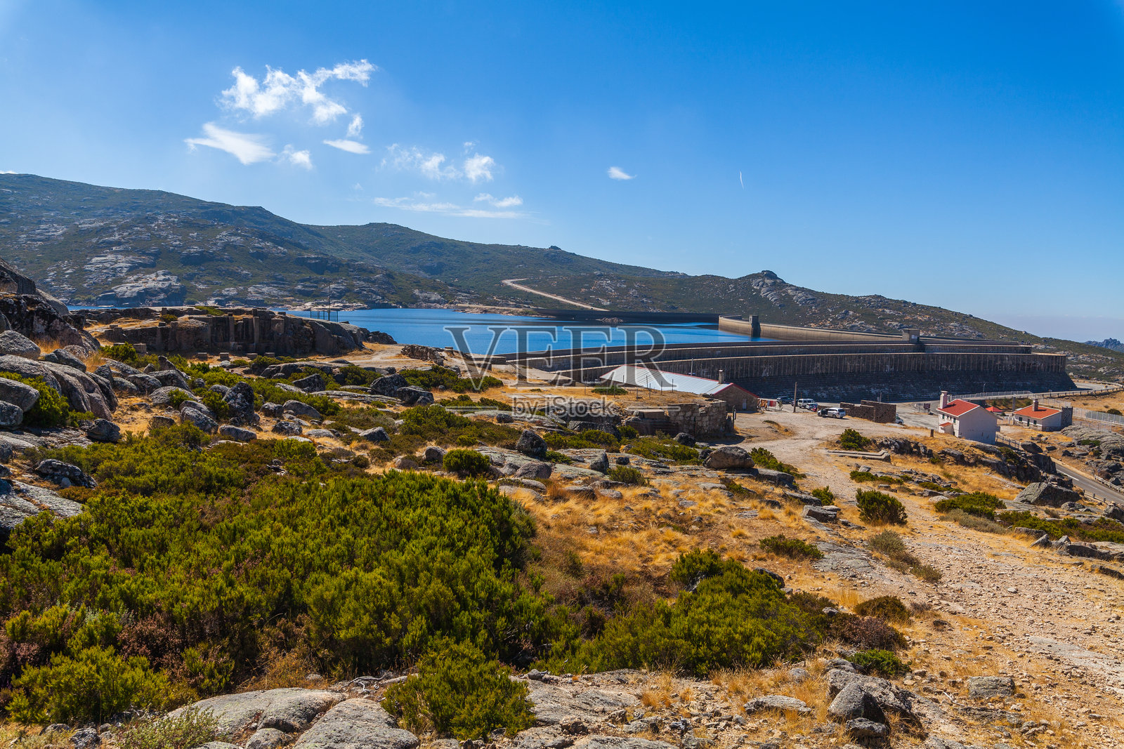 一幅风景画,描绘了一个被山丘和稀疏植被环绕的水库,蓝天上飘着几朵白云。照片摄影图片