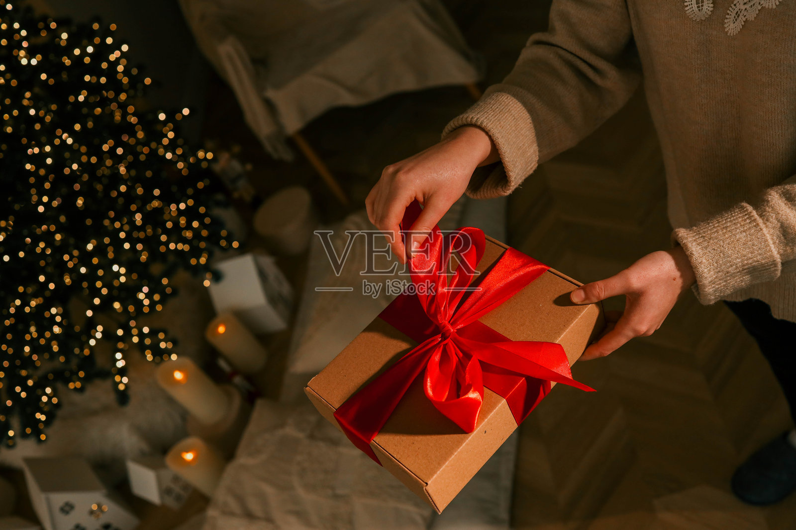 一个人在美丽装饰的圣诞树旁拆开一个系着红色丝带的节日礼物盒。温馨的节日氛围中,背景有靠垫、蜡烛和闪烁的灯饰。照片摄影图片