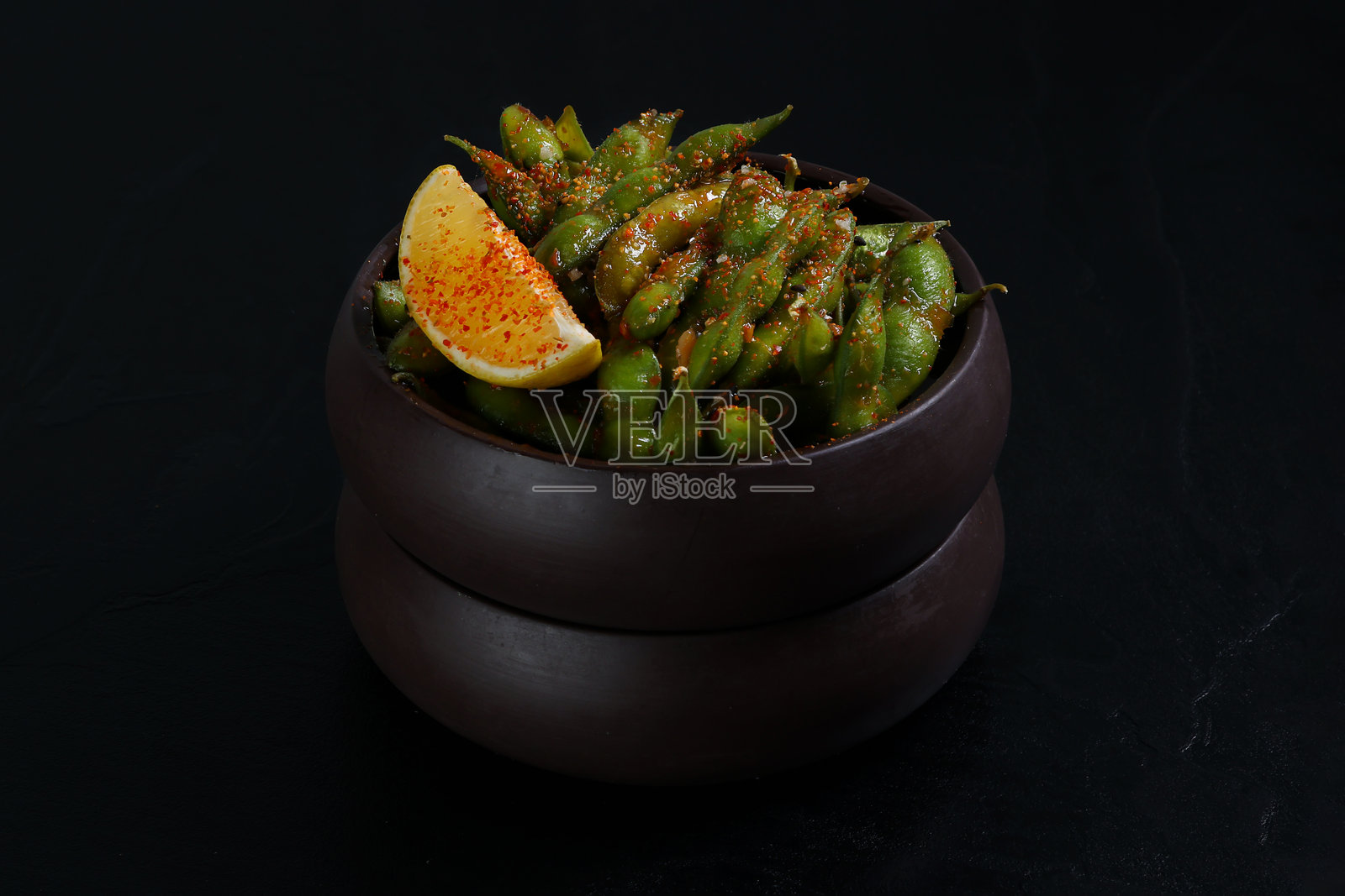 一碗调味的毛豆,配有一片柠檬和撒上辣椒 flakes,呈现出色彩鲜艳且辛辣的小吃,背景为深色。照片摄影图片