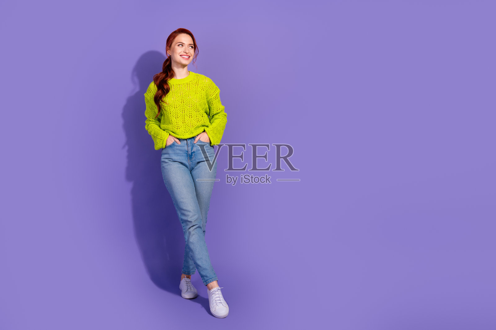 自信的年轻女性穿着明亮的绿色毛衣和牛仔裤,背景是生动的紫色,体现了现代风格和休闲时尚。照片摄影图片