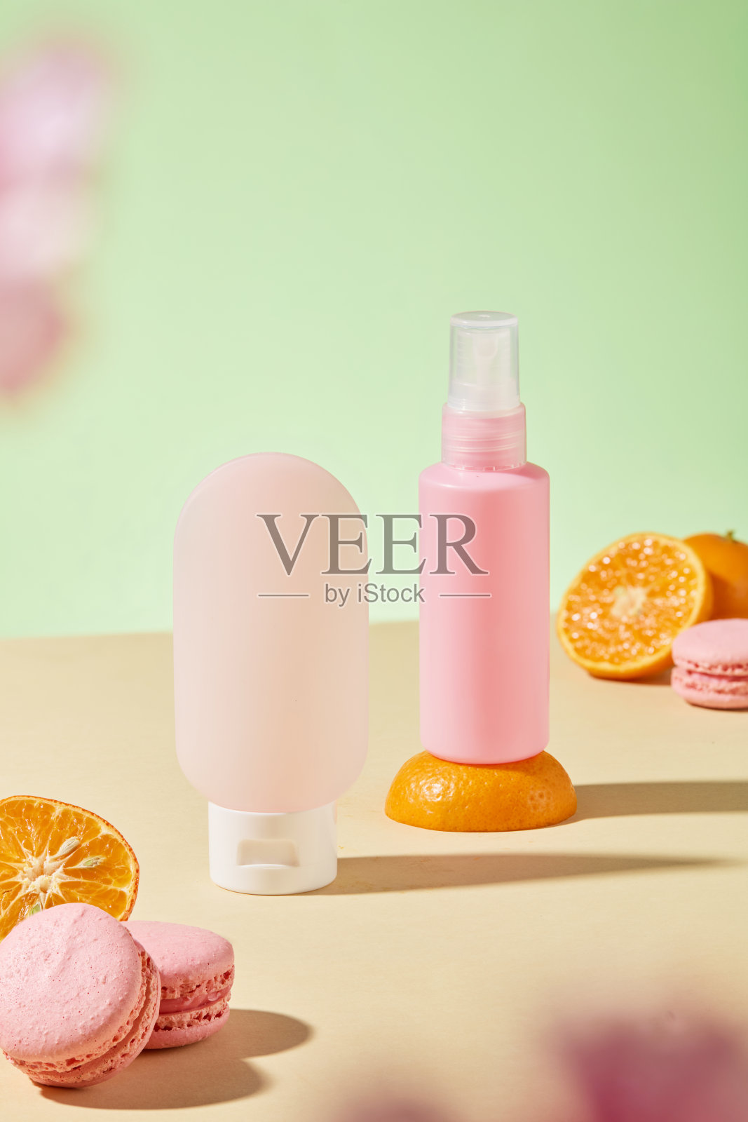 两个无品牌的瓶子在米色背景前展示。瓶子上装饰着切片橙子和粉色马卡龙,背景为绿色。这是一则化妆品广告。照片摄影图片