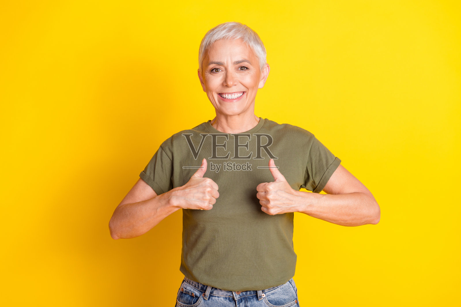 迷人积极的女性穿着卡其色T恤,竖起大拇指,背景为空旷的黄色。照片摄影图片