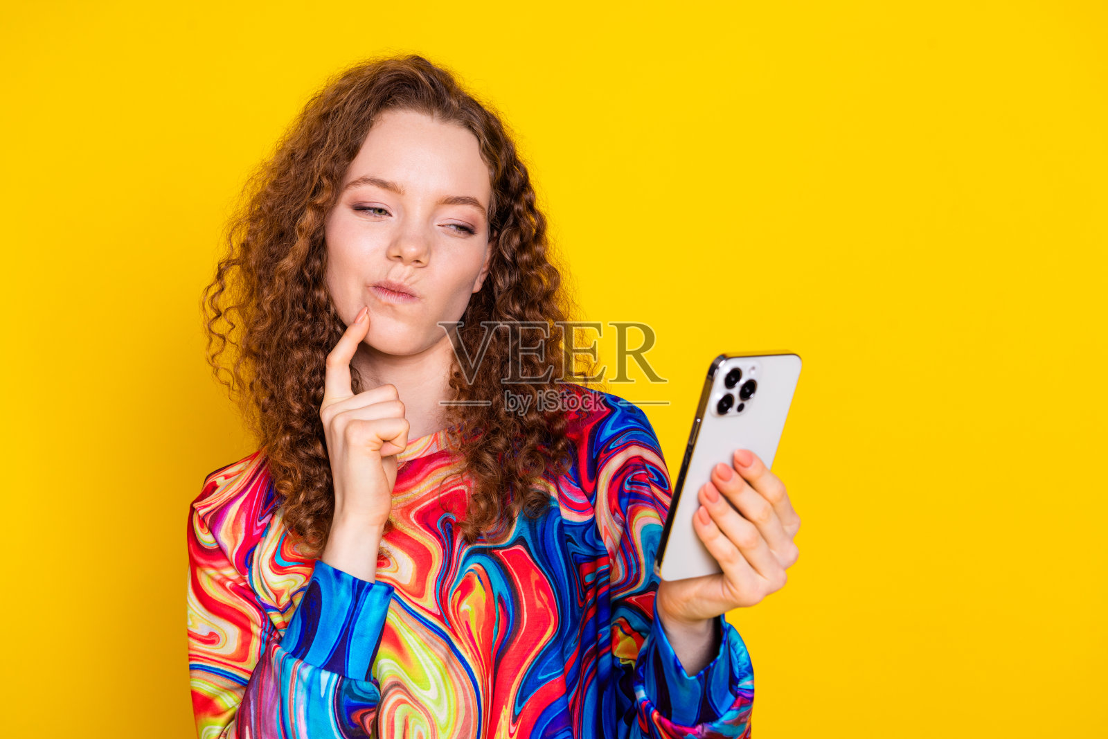 年轻女性穿着时尚印花毛衣,面带沉思表情,手持智能手机,背景为黄色。照片摄影图片