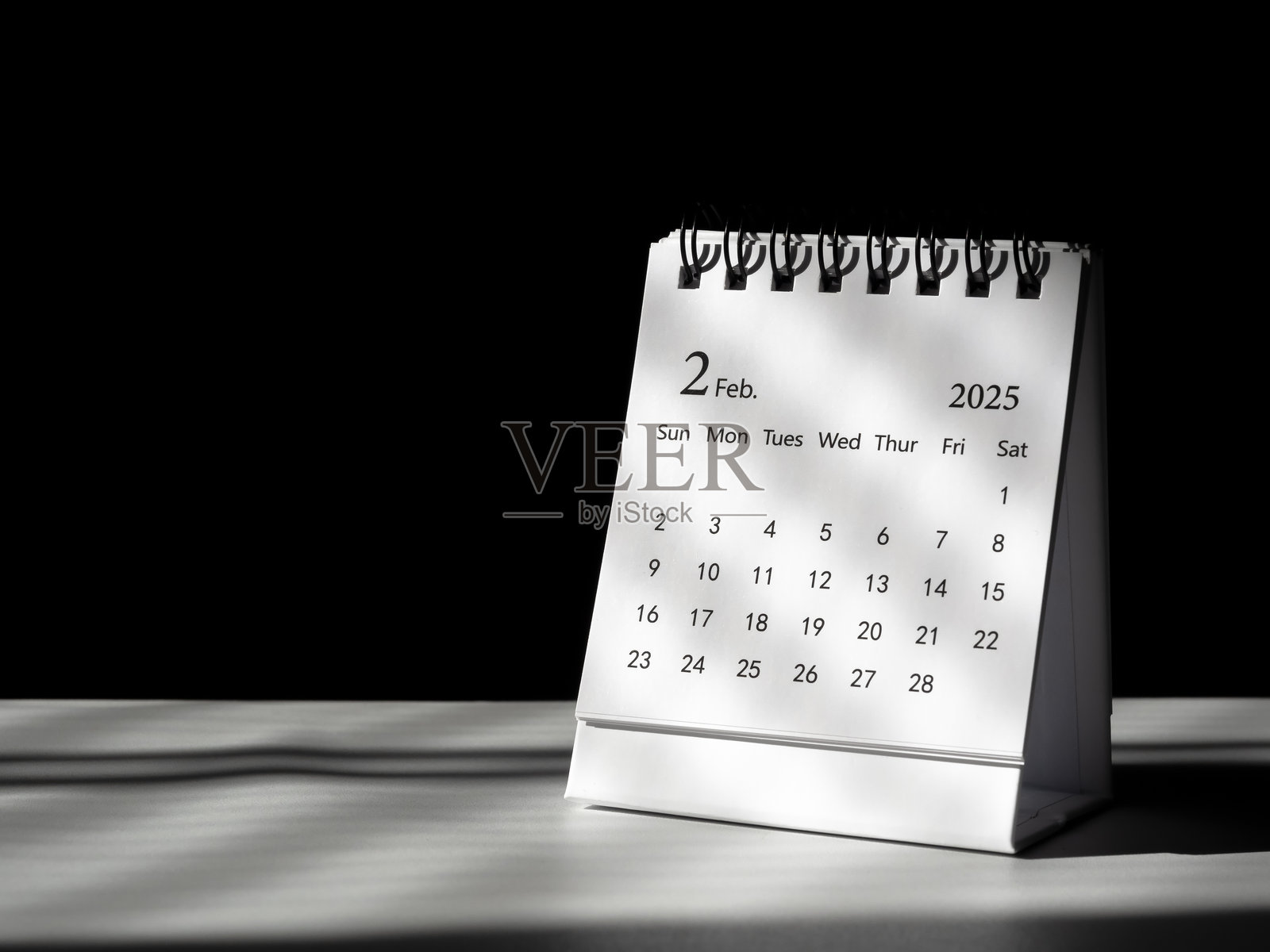 2025年2月的桌面日历,放置在白色桌子上,背景为深色,留有空白空间。小日历显示第二个月的页面,祝您2025新年快乐。照片摄影图片