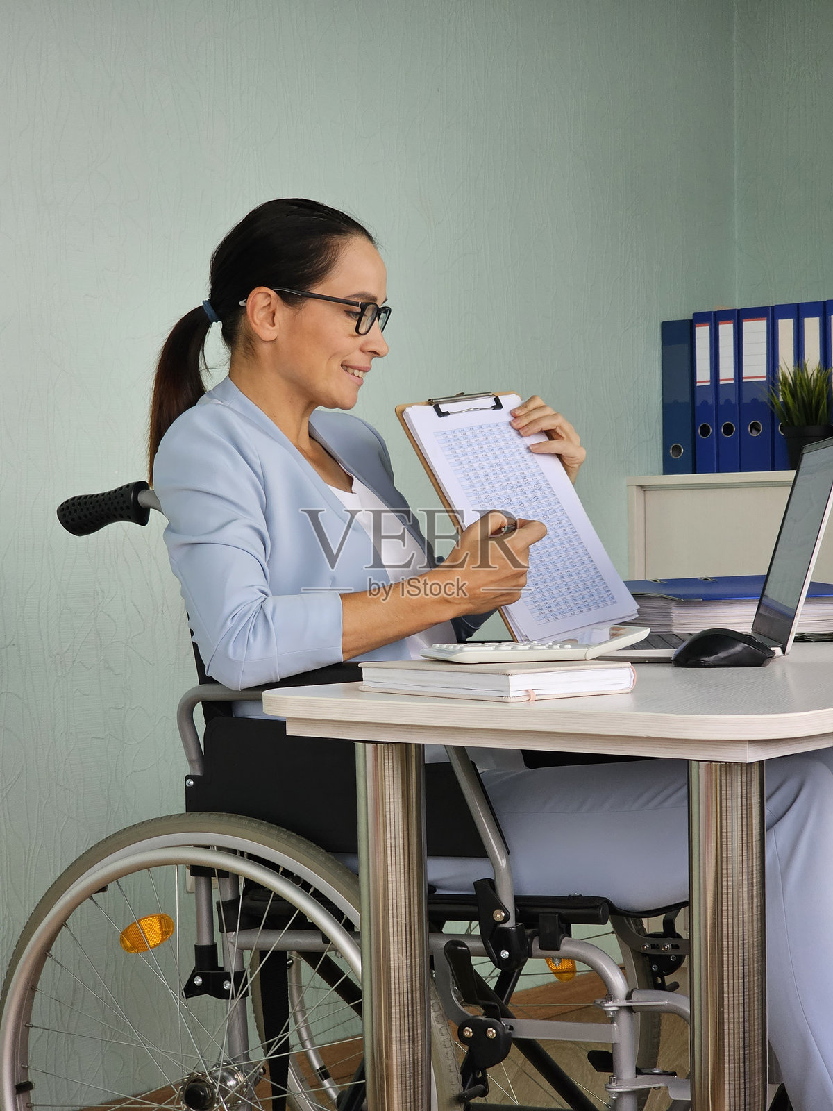 年轻的残疾女教师坐在轮椅上,通过电脑在线咨询学生,并在笔记本上解释笔记。照片摄影图片