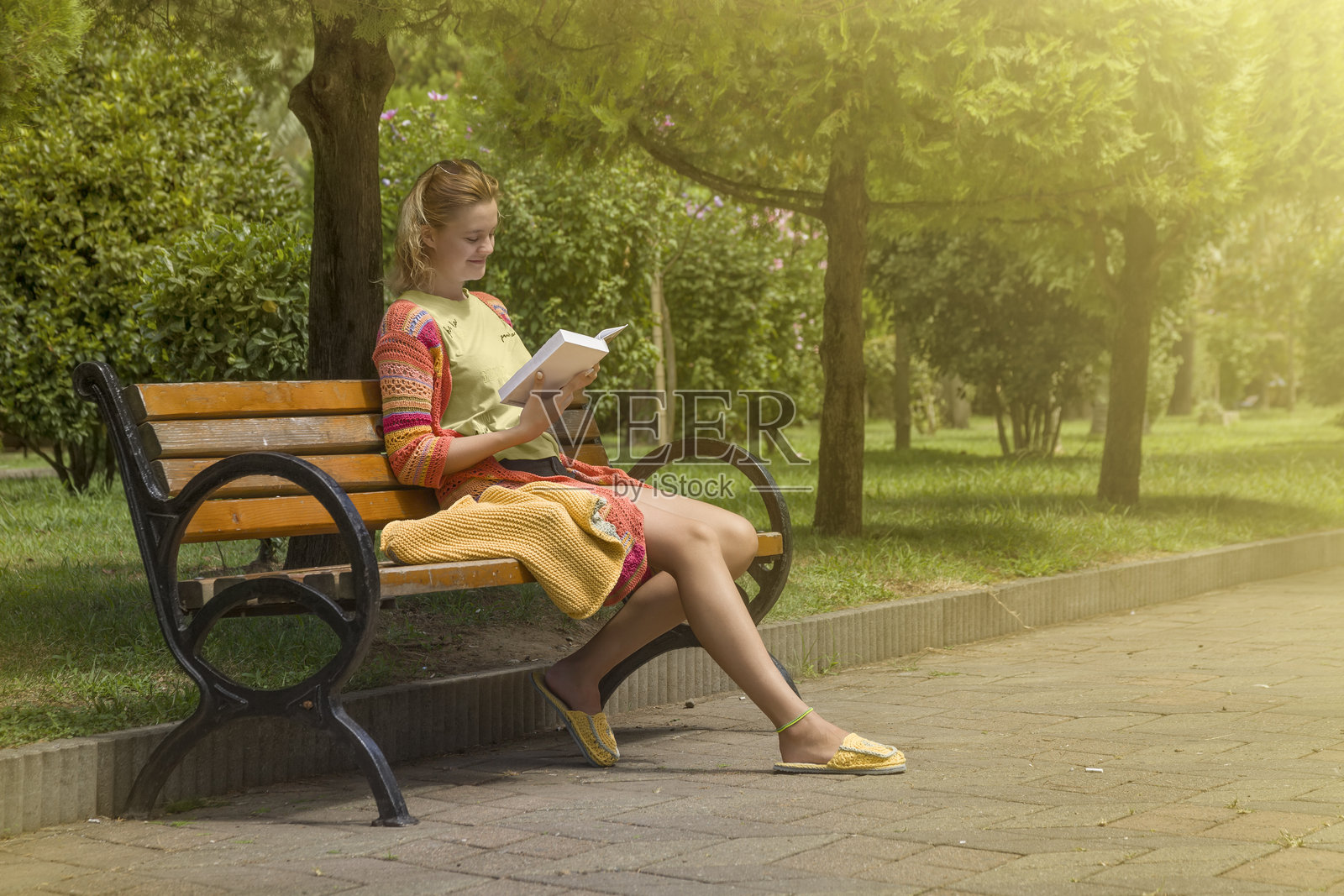 一个穿着棉质针织衣服的小女孩坐在城市公园的木长椅上,享受着温暖的夏日,专心阅读一本书。照片摄影图片
