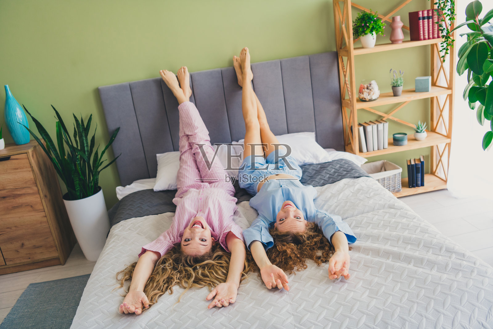 两位年轻漂亮的女性躺在床上,抬起腿,穿着睡衣,醒来时的现代舒适卧室室内光线明亮。照片摄影图片