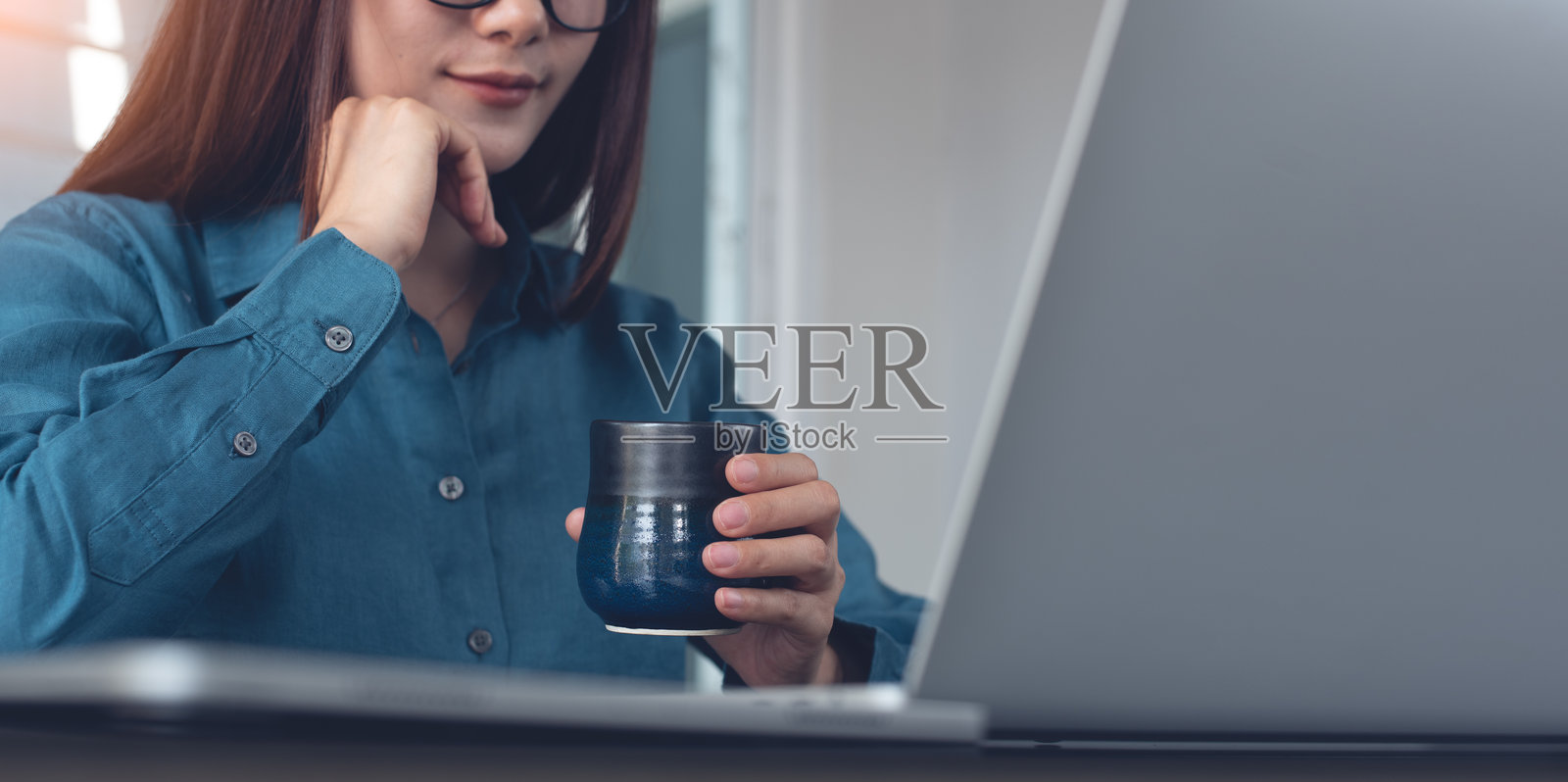 年轻聪明的女商人在现代办公室使用笔记本电脑,手里拿着一杯咖啡,专注地看着屏幕。亚洲女性在在线工作期间享受咖啡休息,同时在笔记本电脑上寻找商业创意。照片摄影图片