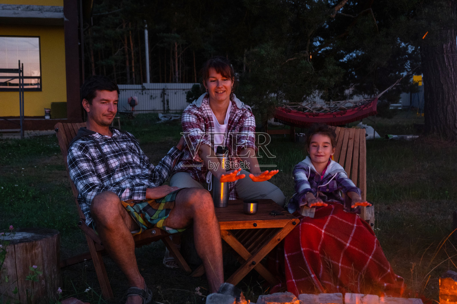 一家人坐在院子里的户外篝火旁,坐在露营椅上,喝着保温瓶里的茶,围着火炉取暖聊天。照片摄影图片