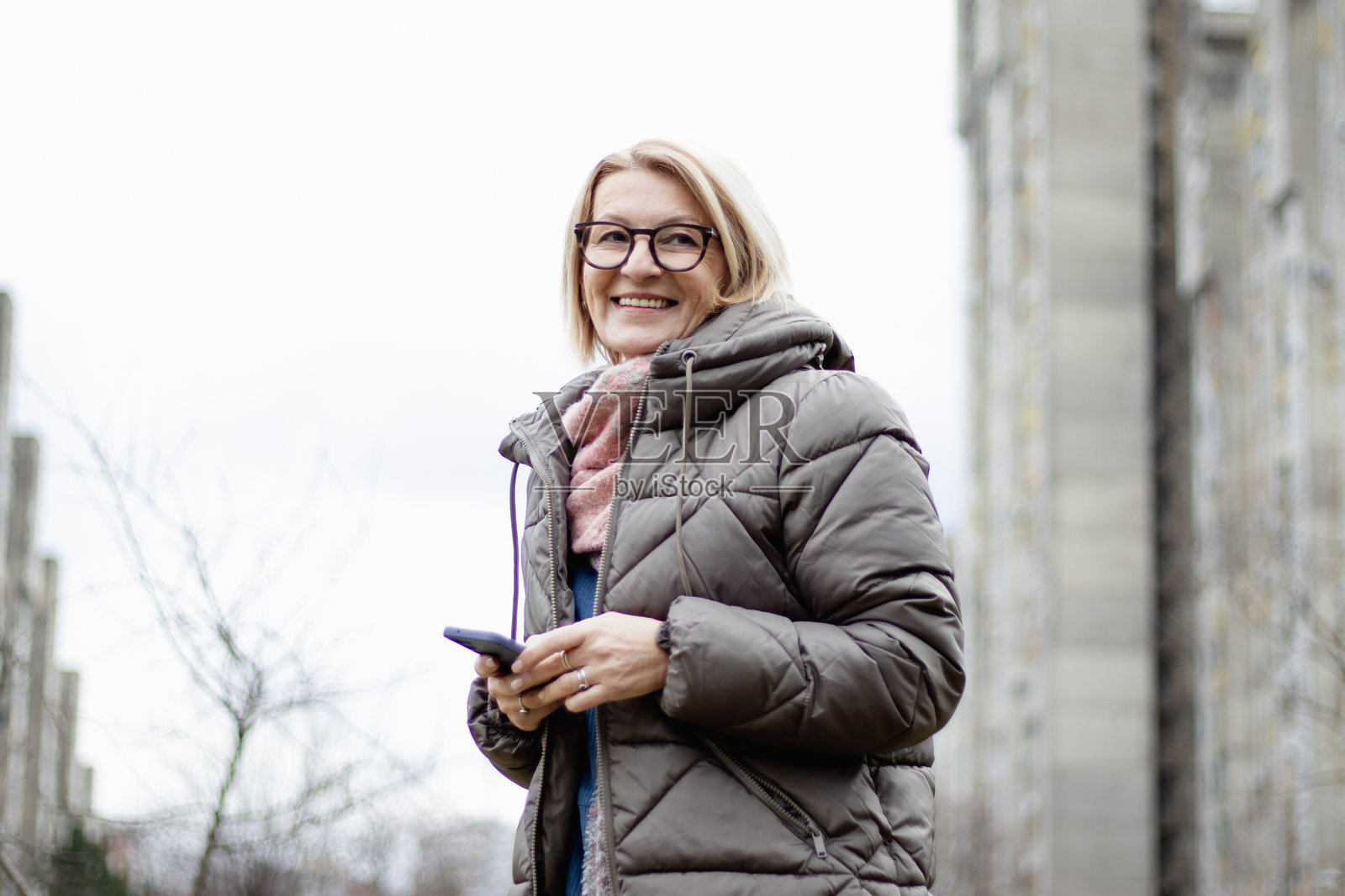 一位成熟的金发开朗女性在寒冷的冬日城市户外戴着眼镜,使用手机。照片摄影图片