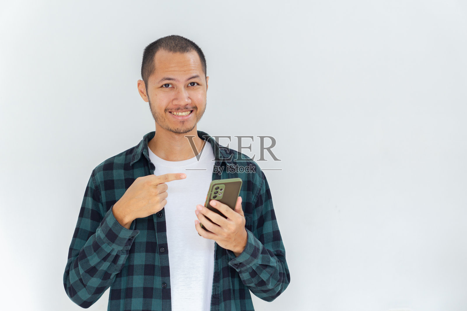 年轻的亚洲男子穿着法兰绒衬衫,面带愉悦表情,指着智能手机,背景为纯白色。照片摄影图片