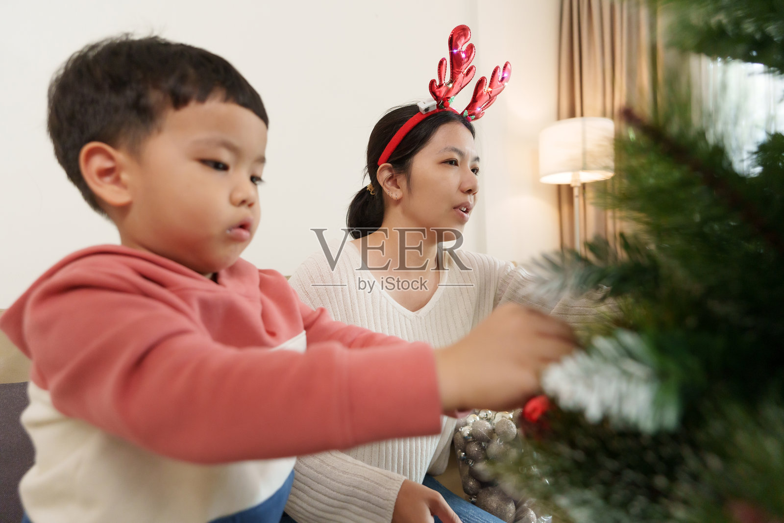 亚洲女性和一个小男孩在温暖的客厅里快乐地装饰圣诞树。孩子在放置装饰品,而女性在协助。气氛非常节日。照片摄影图片