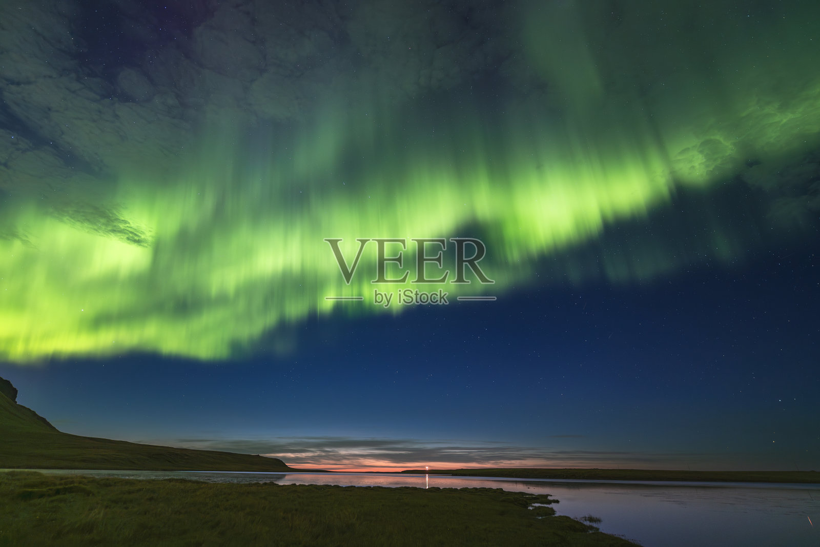 绿色的北极光在黄昏的天空中舞动,湖面上方残留着日落的余晖,位于冰岛西部的格伦达菲厄泽边缘。一个傍晚的童话。照片摄影图片