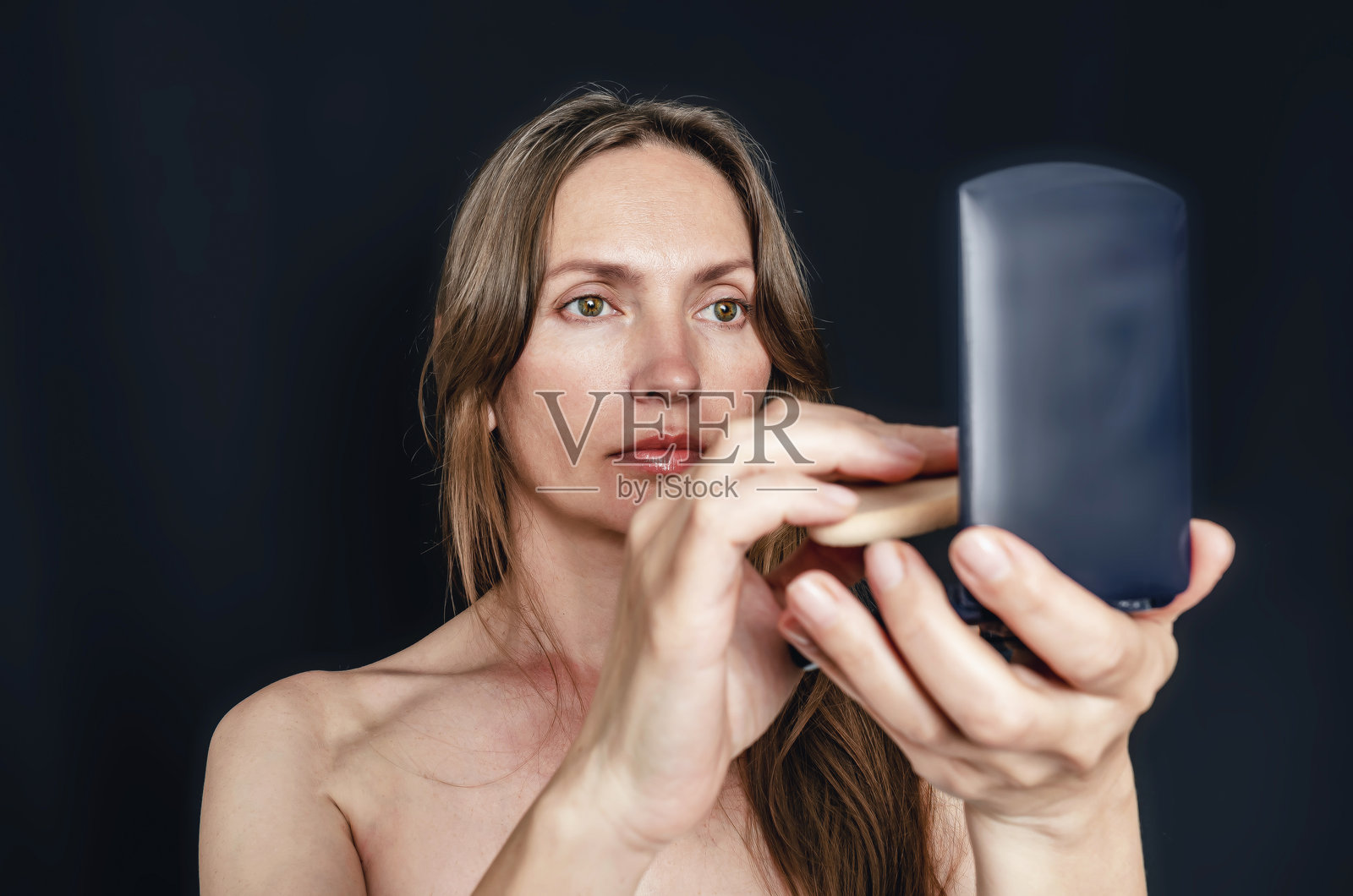 迷人的女性用海绵涂抹粉底,凝视化妆包中的小镜子。照片摄影图片