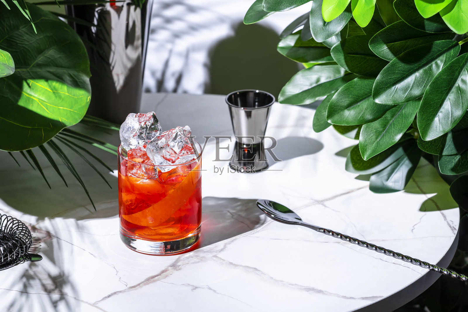 无酒精内格罗尼或纳格罗尼鸡尾酒或无酒精饮品,使用替代金酒、无酒精红味美思和开胃酒。白色大理石桌子上摆放着热带叶子。照片摄影图片