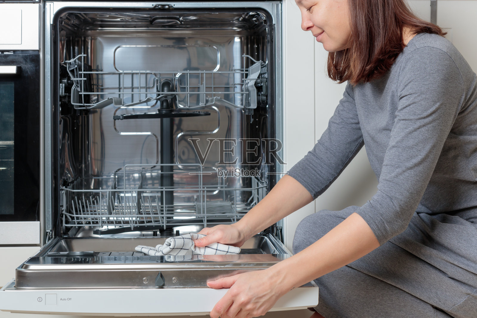 女人用毛巾或抹布清洁打开门的内置空洗碗机。现代住宅的厨房配有简约的白色橱柜。照片摄影图片