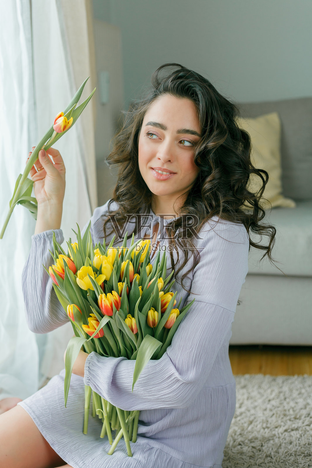 春季肖像:美丽的深色卷发女性穿着紫丁香色连衣裙,手中捧着一大束郁金香花束,置身于家中。情感丰富的模特手握鲜花。国际妇女节。礼物。母亲节。照片摄影图片