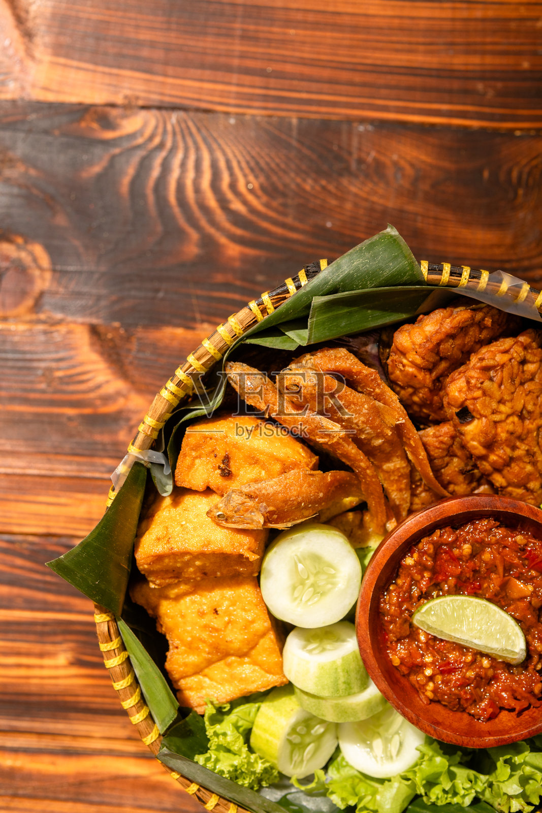 用编织竹篮盛装的传统印尼美食拼盘,包含炸鱼、豆腐、黄瓜、生菜和盛在木碗中的沙爹辣椒酱,摆放在香蕉叶上。照片摄影图片