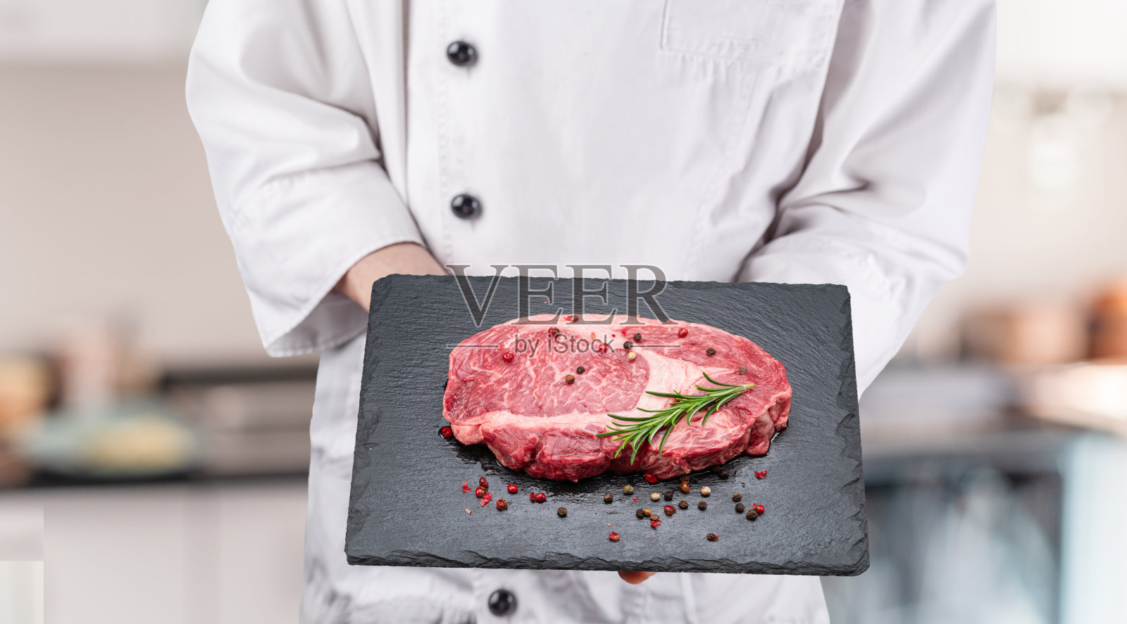厨师手持生肋眼牛排,上面撒有调味料和香草,放在黑色石板餐盘上。照片摄影图片