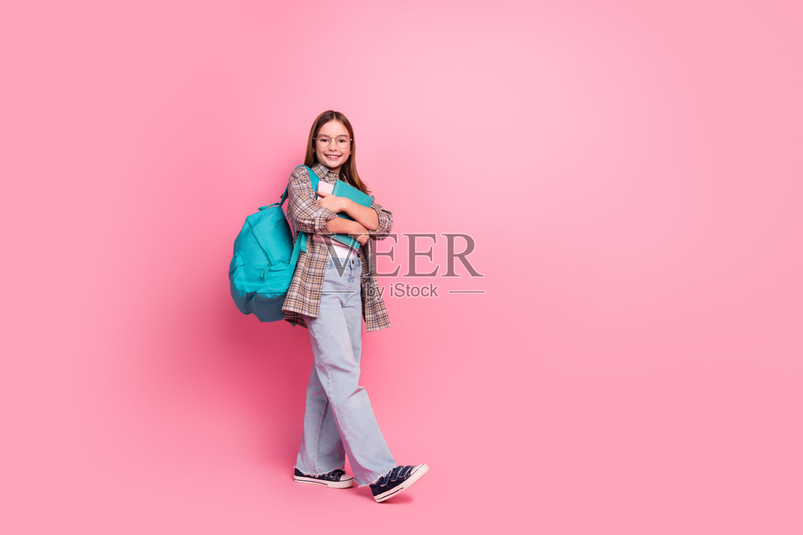 迷人的女学生站在粉色背景前,背着蓝色背包和手拿笔记本,穿着时尚休闲的服装。照片摄影图片