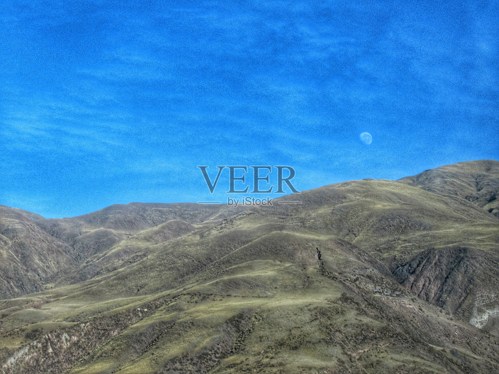 在阿根廷北部萨尔塔省的卡尔查基山谷,壮丽的月亮在蓝天中闪耀,四周被安第斯山脉的巨大多彩山峦环绕,阳光明媚。照片摄影图片