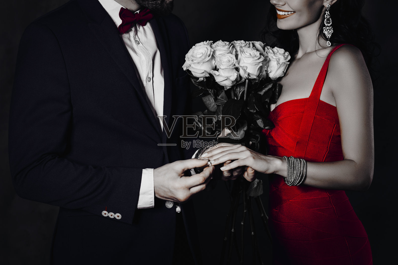 身穿燕尾服的富有绅士为女士戴上镶有钻石的金戒指,女士手中捧着白玫瑰,夫妻二人背景为灰色。照片摄影图片