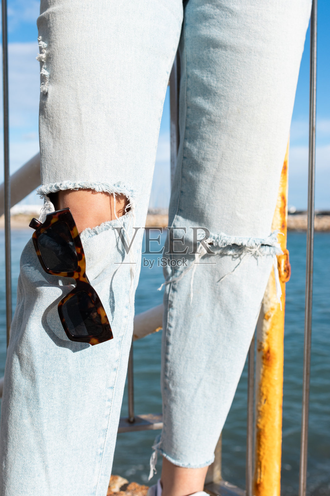 女性穿着膝盖破洞牛仔裤的腿部特写,配有夏季日光浴用的太阳镜。照片摄影图片